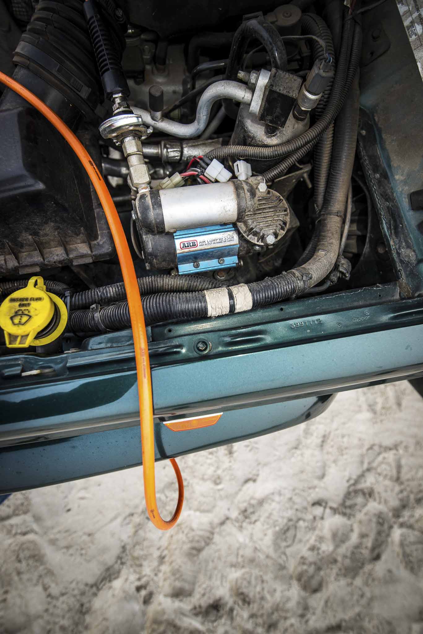 Open car hood showing an ARB air compressor mounted in the engine bay with an orange air line connected to fittings and hoses.