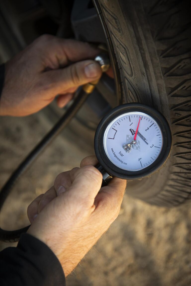 Person inflating/checking a car tire with a pressure gauge attached to the valve stem, readout showing pressure in bar per dial gauge