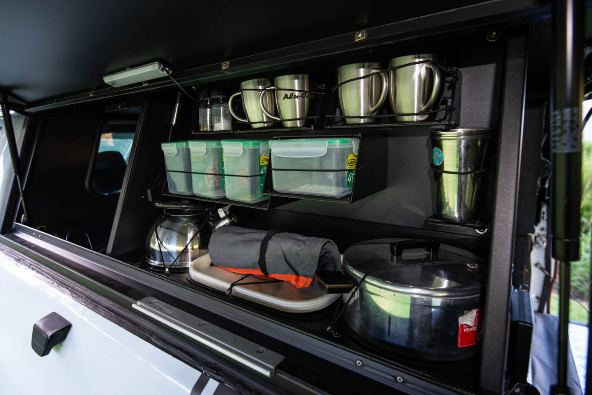 Rear vehicle storage with organized camping gear: mugs, plastic containers, kettle, pots, plates, and a rolled jacket/gear bag inside a drawer system.