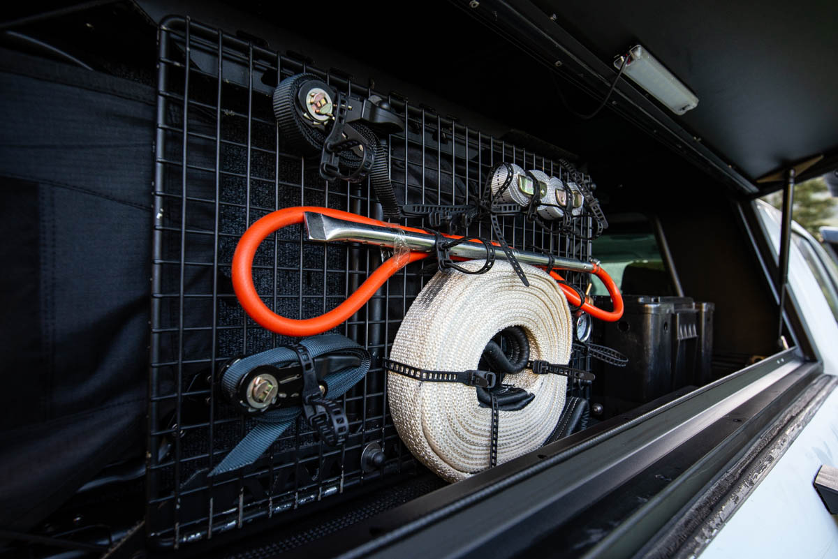 Organized gear in a vehicle trunk: coiled rope, orange rescue bar, and straps mounted on a metal grid panel.