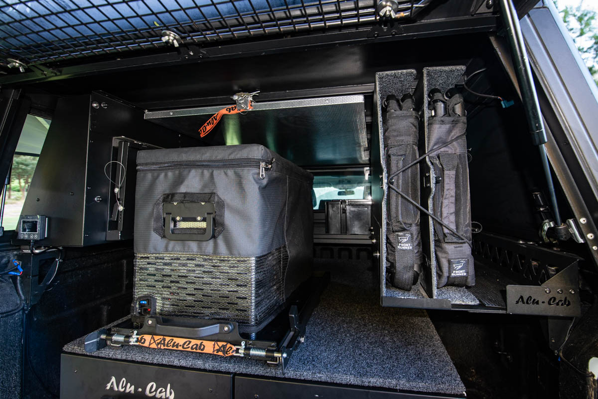 Interior of a vehicle storage area with a large gray gear case, padded racks, and labeled cargo straps secured on a platform.