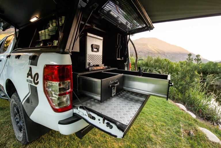 White pickup truck with an open, slide-out metal drawer system in the bed, set up for camping with a diamond-plate platform and toolbox, on a grassy riverside with mountains in the background and sunset light nearby.