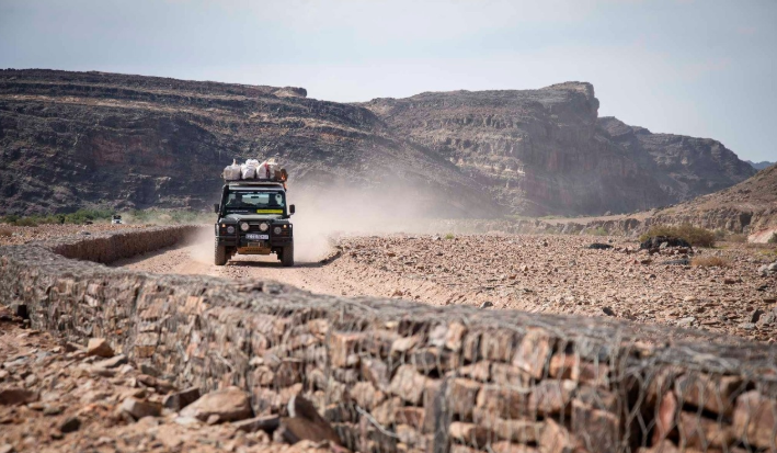 Dark green off-road vehicle kicking up dust on a rocky desert road, with stone walls and bare cliffs in the distance.