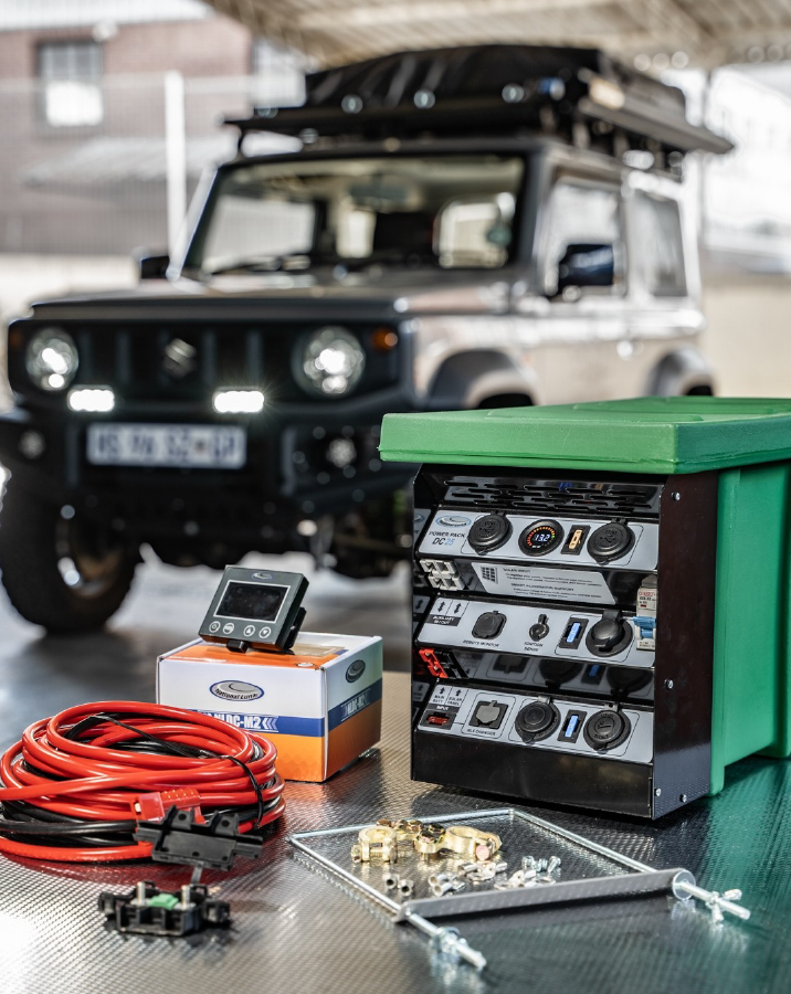 Garage scene: a dark SUV with roof rack in the background; foreground shows a green tool cabinet and automotive electrical components on a metal workbench (red cables, control unit, screws).